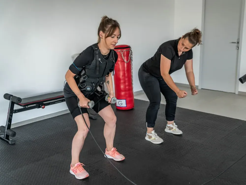 Cliente en combinaison d'électrostimulation guidée par sa coach Get Your Move pendant un exercice de tirage